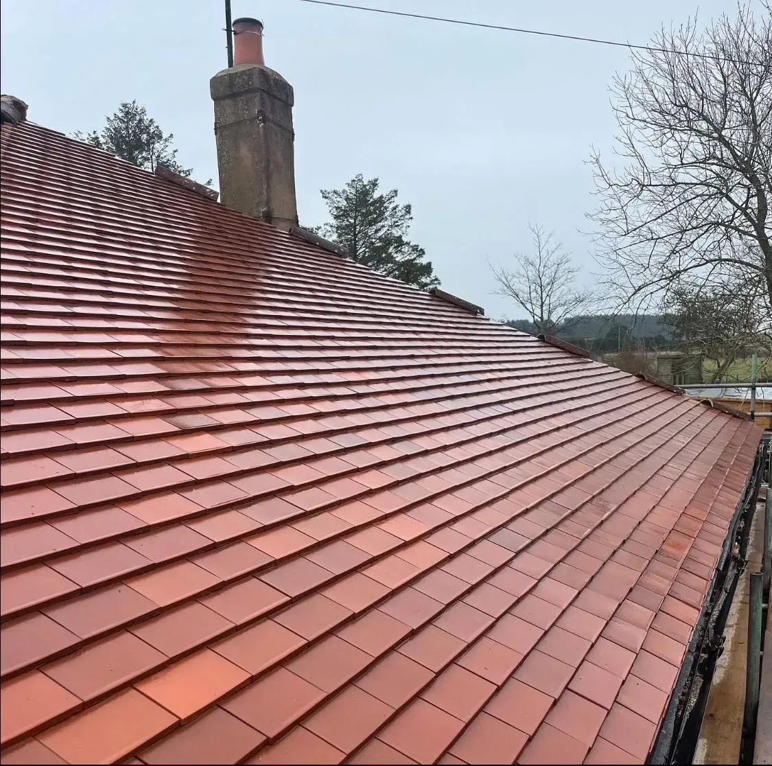 A close-up view of a recently completed pitched roof installation, showcasing new terracotta-coloured plain tiles and a traditional brick chimney stack against a cloudy sky.
