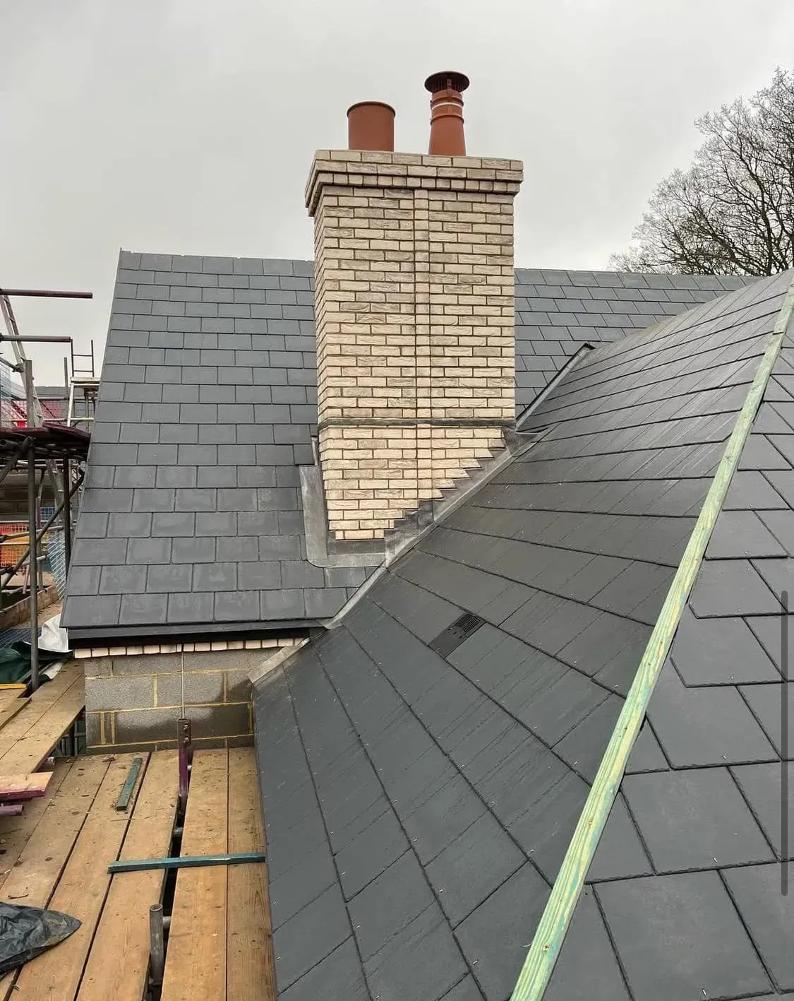 Newly installed dark grey slate roof with a new brick chimney stack and scaffolding.